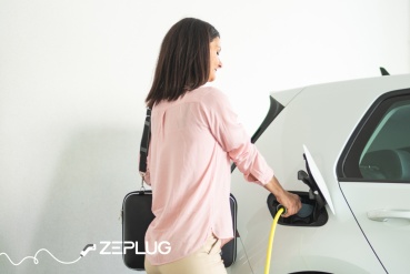 Femme branchant son câble de recharge à sa voiture électrique, tenant un sac de travail, dans un cadre lumineux et moderne.
